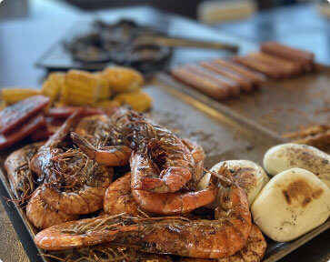 A close-up of a tray of grilled food, with a pile of large shrimp in the foreground.