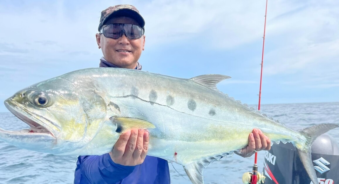 A smiling man on a boat holds up a large silver fish he has caught.