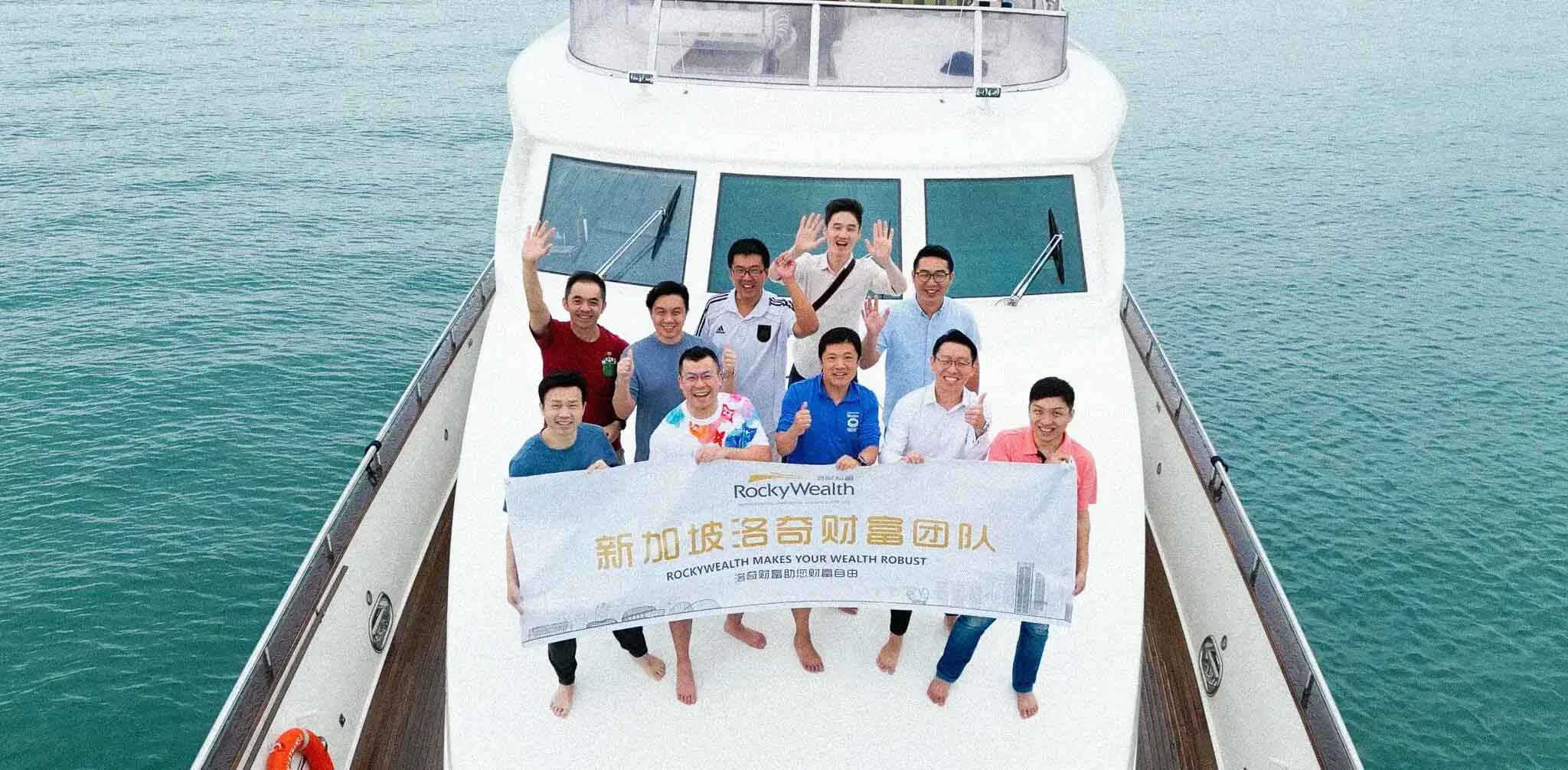 Group of men smiles and waves on the bow of a yacht, holding a large white banner.
