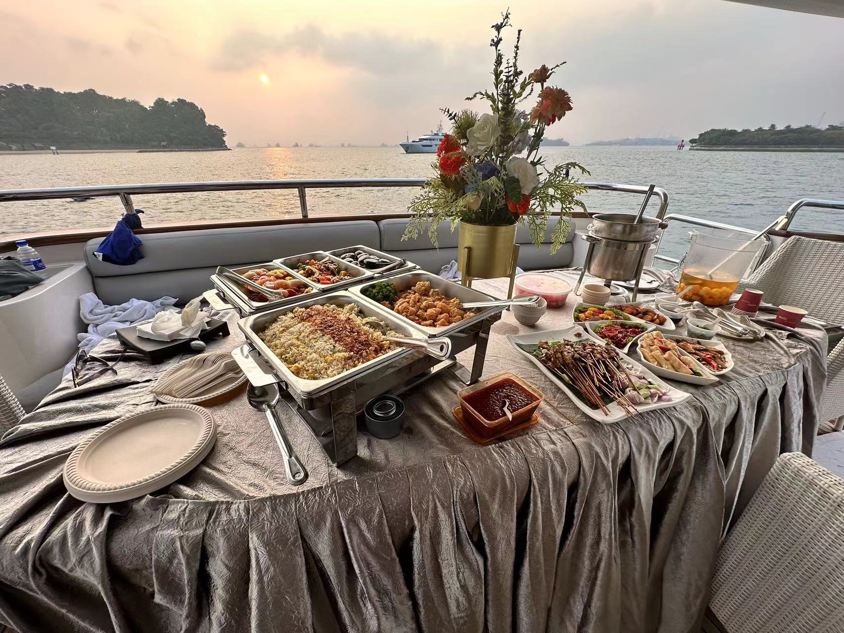 A catered buffet on a yacht deck with chafing dishes, skewers, and flowers, set against a sunset over the sea.
