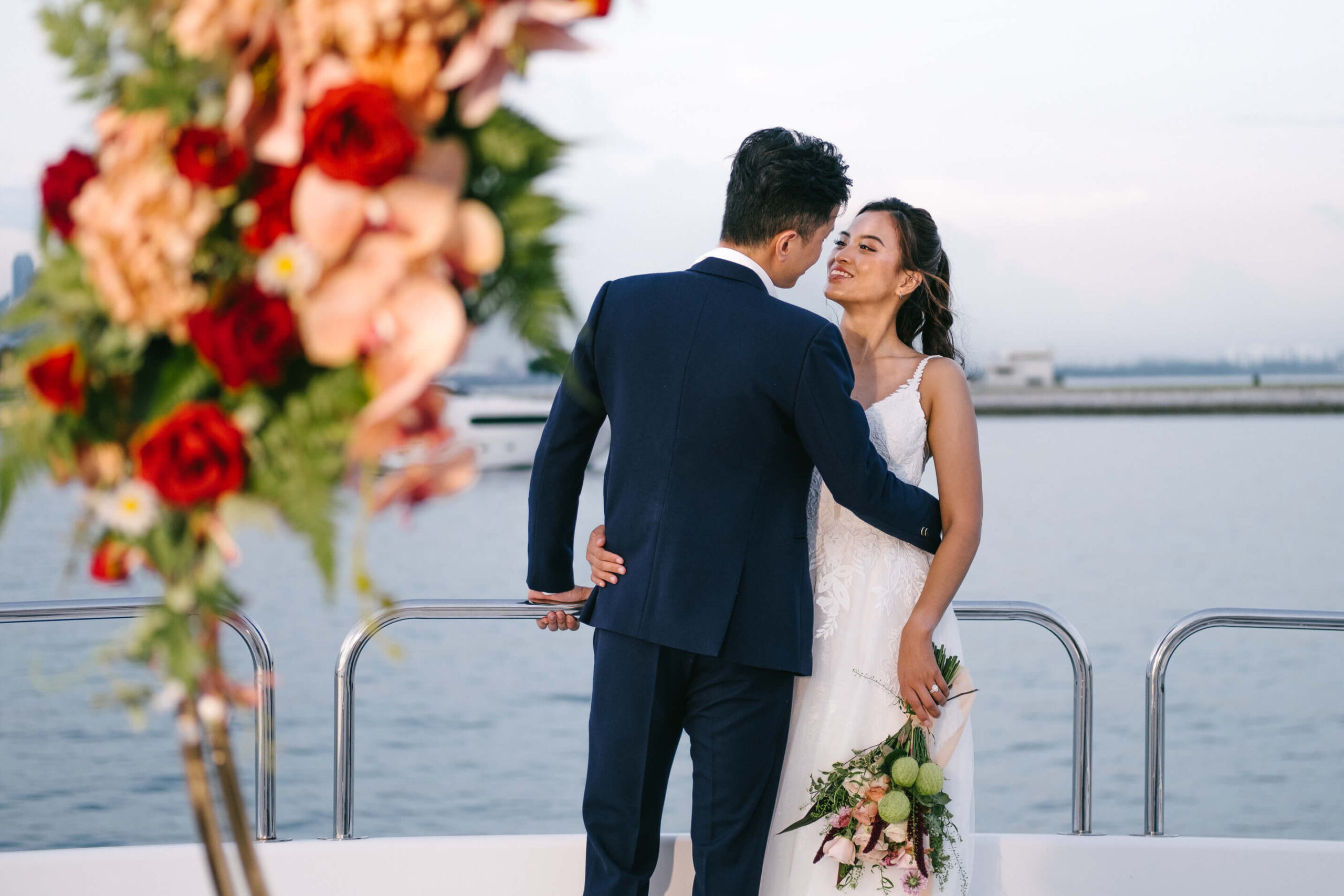 A bride and groom embrace on the deck of a yacht, smiling at each other.